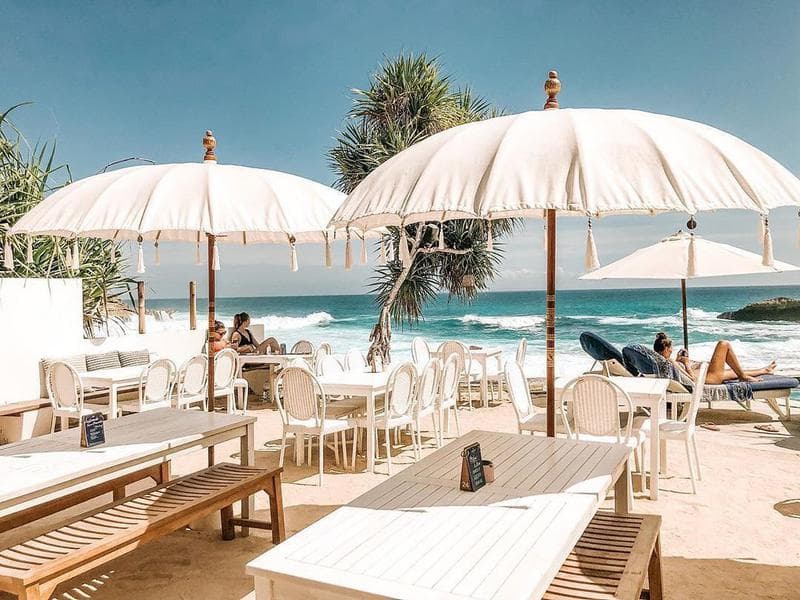 White parasols and tables at the Sandy Bay beach club, with surf breaking on the bay behind